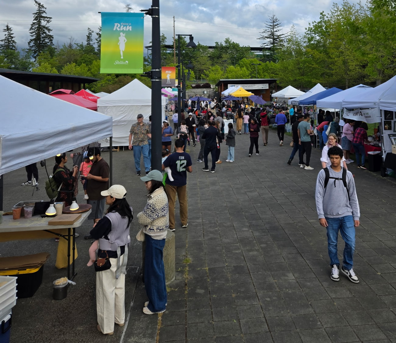 An image depicts a bustling street fair. A large crowd of people is gathered on the sidewalks and street, mingling and enjoying the festivities. There are numerous booths with vendors selling various items. The street itself is lined with parked cars and a food truck can be spotted in the distance. The atmosphere appears lively and vibrant, suggesting a community event. The sky overhead is clear, indicating good weather for this outdoor event.